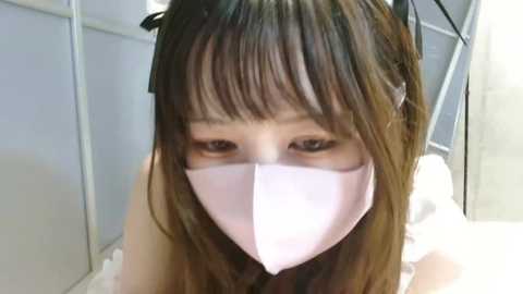 Media: Video of a young Asian woman with long brown hair, wearing a white face mask, gazing intently into the camera, in a sterile medical setting.