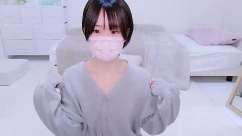 Media: Video of an Asian woman with short black hair and pale skin, wearing a gray cardigan, white mask, and gloves, standing in a minimalist, white bedroom with a bed and dresser.