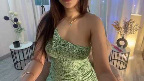 Media: Video of a young woman with long dark hair, wearing a green and black polka-dot one-shoulder dress, sitting on a light-colored couch in a modern, softly lit room with white curtains, minimalist decor, and floral arrangements.