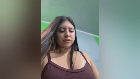 Media: A video of a Latina woman with long black hair, wearing a low-cut, maroon tank top, adjusting her hair in a bright, modern room with green and white walls.