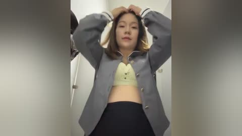 Media: Video of an East Asian woman in a grey blazer and black skirt, adjusting her hair, standing in a narrow, white-walled hallway.