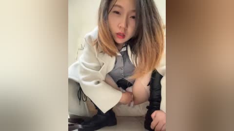 Media: Video of an East Asian woman with long, straight brown hair, wearing a white leather jacket, gray blouse, and black pants, squatting provocatively in a neutral-toned room.