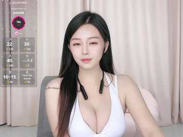 Media: A video of an Asian woman with long black hair, fair skin, and large breasts in a white tank top, sitting in a living room with beige curtains.