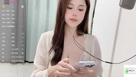 Media: Video of an Asian woman with long brown hair, wearing a white cardigan, listening to headphones while holding a small device, in a clean, bright room with a gray scale wall chart in the background.