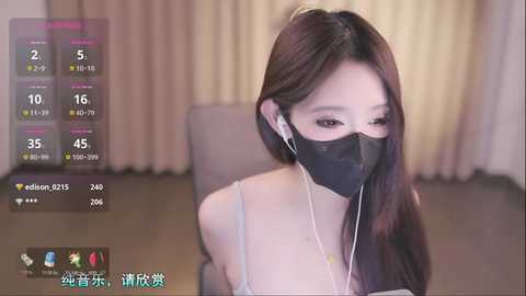 Media: Video of an East Asian woman with long brown hair, wearing a black face mask, white headphones, and a light blue spaghetti strap top, sitting indoors with beige curtains and a gray chair in the background.