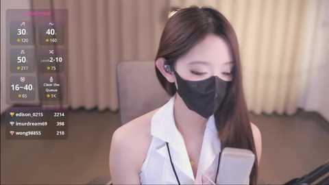 Media: Video of an East Asian woman with long, straight brown hair, wearing a black mask, white blouse, and headphones, seated in a modern office.