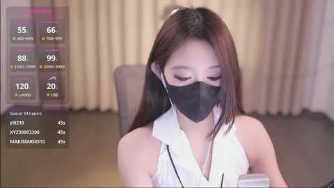 Media: Video of an East Asian woman with long, straight dark hair, wearing a black face mask and a sleeveless white top, seated indoors. The background features beige curtains and a gray chair.