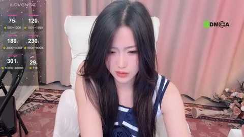 Media: Video of a young East Asian woman with long, dark hair, wearing a navy blue dress, sitting on a white chair in a living room with beige curtains.