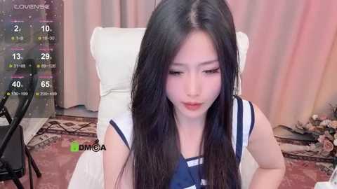 Media: A video of an Asian woman with long black hair, wearing a white sleeveless top, sitting on a white chair, in a room with a red patterned rug, pink curtains, and a black chair.