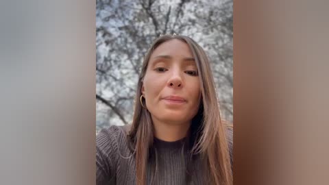 Media: A video of a young woman with light brown skin, straight brown hair, and a neutral expression. She wears a grey ribbed sweater. The background features blurred tree branches.