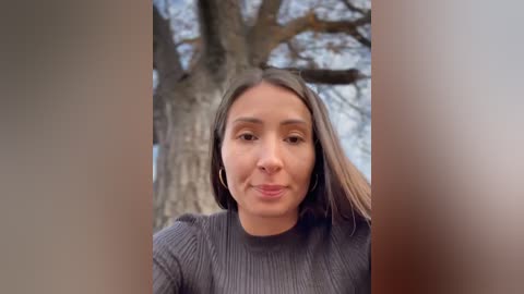 Media: Video of a young woman with light brown skin, long dark hair, and hoop earrings, wearing a dark sweater, standing in front of a large tree trunk in a blurred outdoor setting.