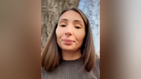 Media: Video of a young woman with straight, shoulder-length brown hair, wearing a grey ribbed sweater, making a pouty face. Background features a blurry, textured stone wall.