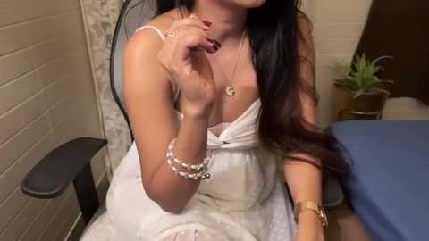 Media: Video of a woman with long dark hair, wearing a white dress, sitting in a chair, holding her finger to her lips, surrounded by a brick wall and a potted plant.