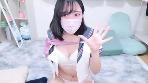 Media: Video of an East Asian woman with straight black hair, wearing a pink face mask, white lace bra, and a purple and white schoolgirl outfit, waving in a softly lit room with pastel furniture.