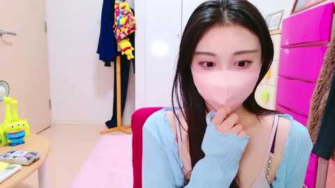 Media: Video of a young Asian woman with long black hair, wearing a light blue shirt, pink face mask, and a pink handbag, standing in a brightly lit room with colorful clothing and toys.