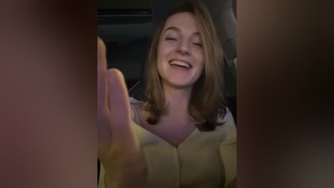 Media: Video of a smiling Caucasian woman with shoulder-length brown hair, wearing a yellow blouse, seated in a dimly lit car. She is reaching out of frame.