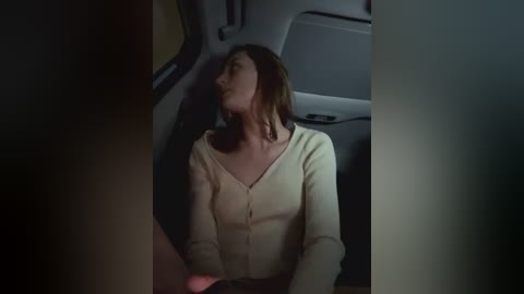 Media: Video of a Caucasian woman with shoulder-length brown hair, wearing a cream-colored, button-up cardigan, sitting in the backseat of a dimly lit car, head tilted slightly back, looking serene.