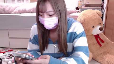 Media: Video of an Asian woman with long brown hair, wearing a face mask, reading a magazine in a cozy bedroom with a teddy bear and a neatly made bed in the background.