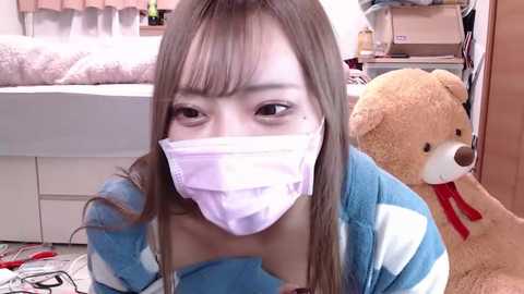 Media: Video of an East Asian woman with long brown hair, wearing a light blue sweater, a white face mask, and a large teddy bear in a cluttered bedroom with a bed and a dresser.