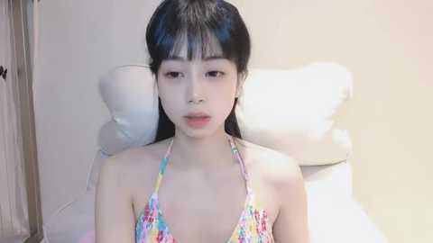 Media: A video of a young East Asian woman with straight black hair and pale skin, wearing a colorful halter bikini top, seated on a white cushion against a plain beige wall.