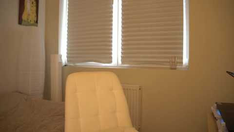 Media: A video of a minimalist bedroom featuring a white upholstered chair, beige bed with a striped bedspread, and closed white blinds. The room has a warm, neutral color palette.