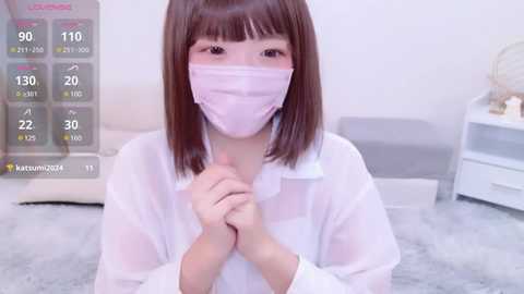 Media: Video of an Asian woman with straight, shoulder-length brown hair, wearing a white mask, white blouse, and grey skirt, kneeling on a plush grey carpet. Background features a white wall and a white dresser with decorative items.