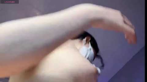 Media: Video of a woman with light skin and shoulder-length black hair, seen from the side. She's stretching her right arm above her head, revealing her bare back and a glimpse of her left breast. The background is a blurred, dark room with purple and blue tones.