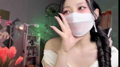 Media: Video of an East Asian woman with long black hair in braids, wearing a white mask, off-shoulder white top, and touching her face, set in a dimly lit room with floral decor and a green light.