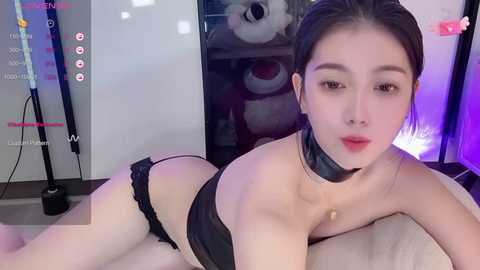 Media: Video of a young Asian woman with fair skin and dark hair, lying provocatively on a bed in black lingerie. Background includes a plush toy and a mirror, with a video game interface overlay.