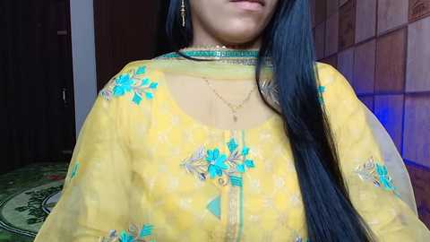 Media: Video of a woman with long black hair, wearing a yellow kurta with turquoise embroidery, and a choker necklace, indoors with a tiled wall and a green rug in the background.