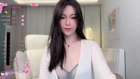 Media: A video of an East Asian woman with long, straight black hair, fair skin, and a slender physique, wearing a sheer white cardigan over a teal lace bra, sitting in a modern, minimalist room with a white chair, beige curtains, and a framed painting on the wall.