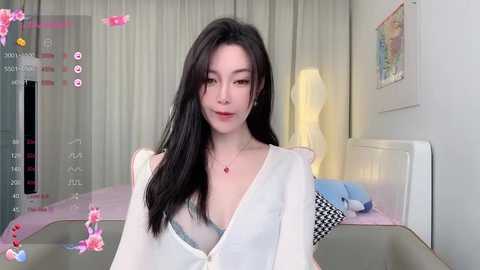 Media: A video of an East Asian woman with long black hair, fair skin, and a slim physique, wearing a white blouse, sitting in a modern bedroom with white curtains, a bed, and a lamp.