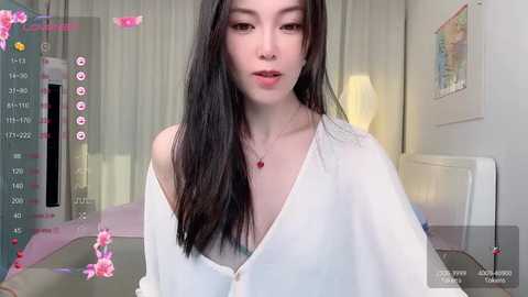 Media: Video of a slender Asian woman with long black hair, wearing a loose white shirt, standing indoors near a window, with a pink flower filter overlay.