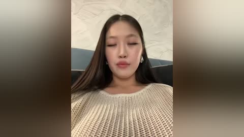 Media: Video of an East Asian woman with straight, dark hair, wearing a beige, textured knit sweater, closing her eyes and pouting. Background shows a textured white wall and a blue object.