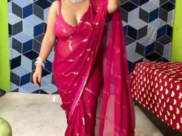 Media: Video of a woman in a vibrant pink saree with sheer fabric, revealing a white blouse underneath. She stands in a room with geometric patterned wallpaper and a red bedspread.