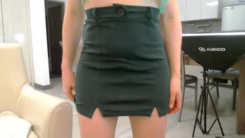 Media: Video of a woman in a teal, high-waisted skirt with a small keyhole cutout at the hem, standing in a modern kitchen with white cabinets, beige sofa, and a Nikon camera on a tripod.