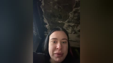 Media: Video of a woman with long black hair and fair skin, puckering her lips, against a dimly lit, textured wall with cracks and patches of peeling paint.