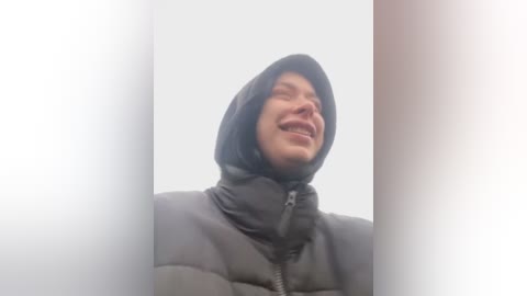 Media: Video of a smiling man in a black puffer jacket, hood up, against a bright, foggy background. His eyes are squinted shut, and his expression is joyful.