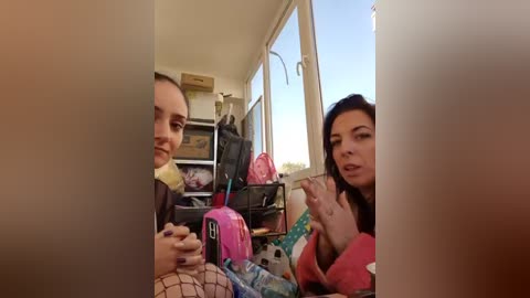 Media: Video of two women, one with short hair and the other with long hair, sitting on a bed with cluttered surroundings, including backpacks and a pink water bottle.