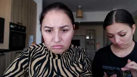 Media: Video of two women: one with zebra-patterned top, frowning, another in black shirt, texting. Modern kitchen background.