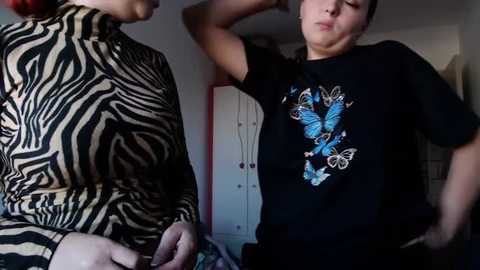 Media: Video of a woman in a zebra-patterned turtleneck and another woman in a black T-shirt with a butterfly design, both in a dimly lit room.