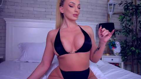 Media: Video of a blonde, curvy woman in a black bikini, sitting on a bed with white sheets. She holds a glass, wearing heavy makeup. Background includes a brick wall, potted plants, and a white headboard.