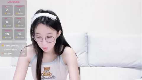 Media: Video of an East Asian woman with long black hair, wearing glasses, a white headband, and a tank top with a teddy bear design. Background features a white couch and a calendar screen with health metrics.