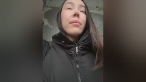 Media: Video of a young Asian woman with long black hair, wearing a black puffy jacket, smiling with eyes closed, standing in a dimly lit room with a textured ceiling.