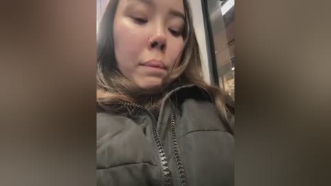 Media: A close-up video of a young woman with fair skin and light brown hair, wearing a dark green puffy jacket, looking down thoughtfully. Background is blurry and indoor.