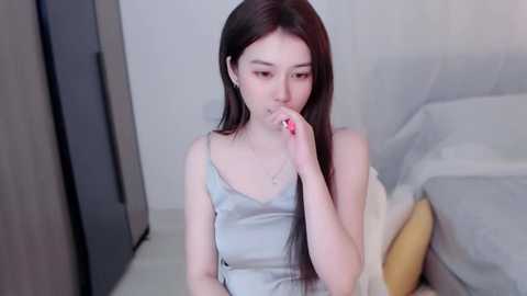 Media: Video of an East Asian woman with long black hair, fair skin, and small breasts, wearing a light blue camisole, sitting on a bed in a modern, minimalist bedroom. She is covering her mouth with her right hand.