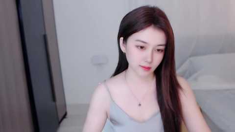 Media: A video of a young Asian woman with long, straight black hair, wearing a light grey spaghetti strap tank top, sitting on a bed in a modern, minimalist bedroom with white walls and grey bedding.