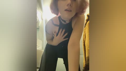 Media: Video of a young woman with short pink hair, wearing a black halter top, leaning against a wall in a dimly lit bathroom, hand on chest.