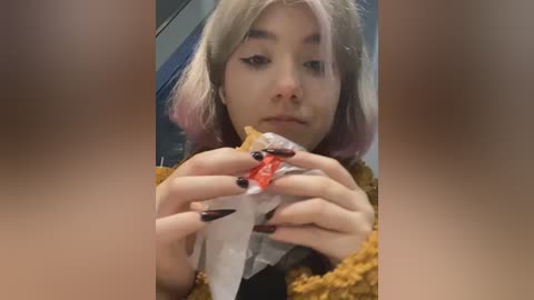 Media: Video of a young woman with pastel pink hair, wearing black nail polish, eating a burger with a red sauce. She's dressed in a mustard-yellow sweater.