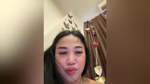 Media: Video of an Asian woman with long black hair, wearing a headband adorned with butterflies, in a bedroom with beige walls and brown curtains, giving a playful kiss to the camera.